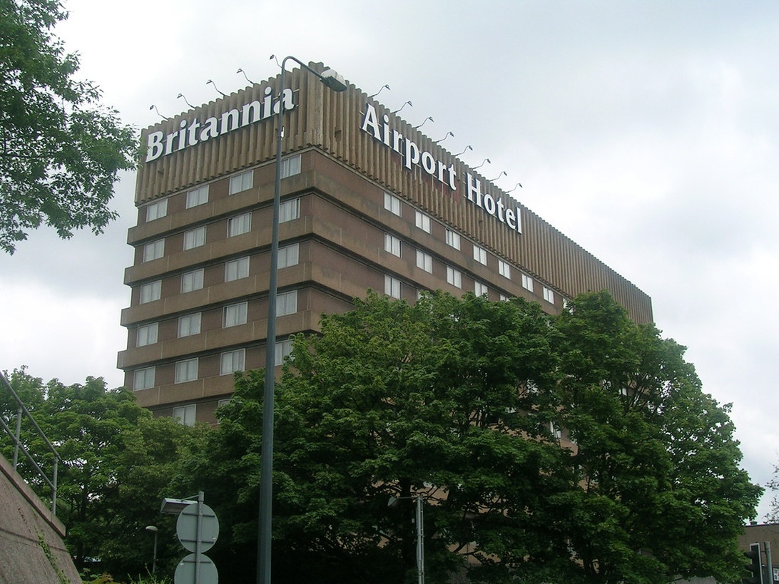 Das Äußere eines mehrstöckigen Flughafenhotels, des 'Britannia Airport Hotel', mit Bäumen im Vordergrund, was auf eine gewisse Begrünung trotz Flughafennähe hindeutet.